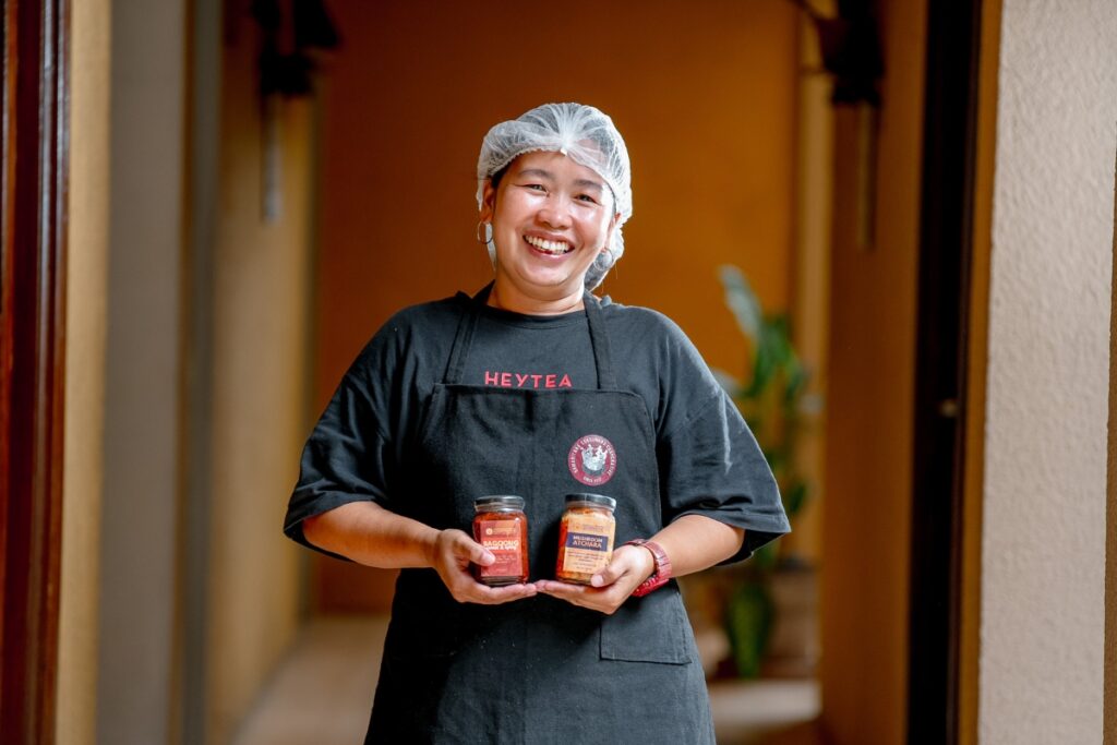 a woman with a black apron and hair net holds two jars of sauces