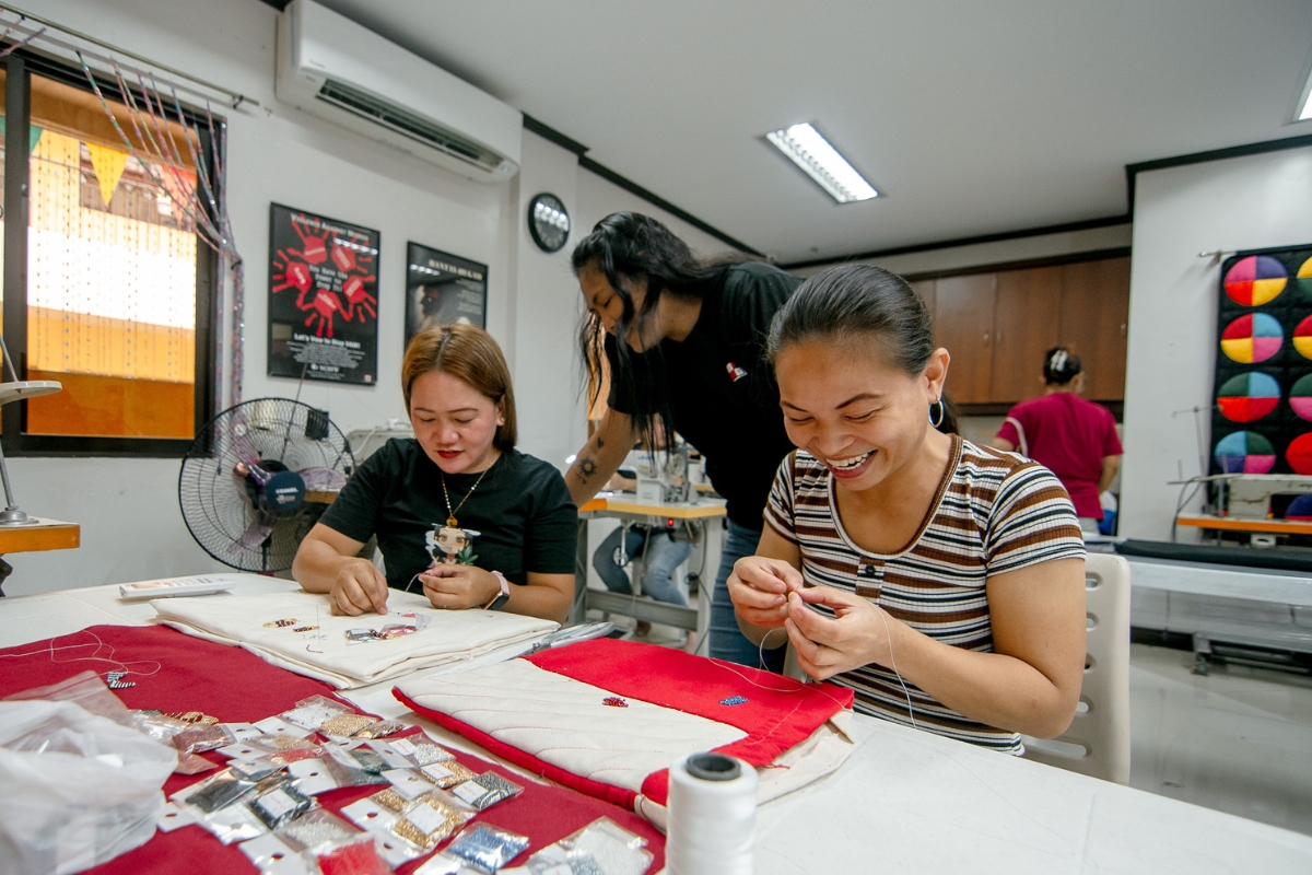 two women sit at a table working with beads and thread