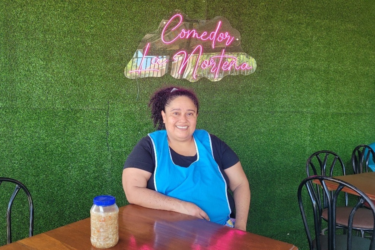 a woman sits at a table in front of a sign saying "Comedor La Norteña"