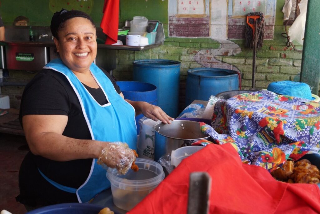 a woman in a blue apron smiles with a container of food and a pot in front of her