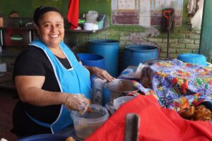 a woman in a blue apron smiles with a container of food and a pot in front of her