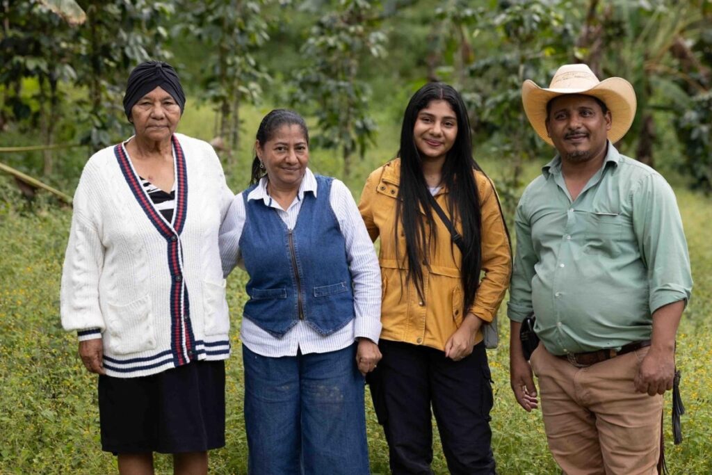 an old woman, a middle-aged woman, a young woman, and a middle-aged man stand smiling in front of vegetation