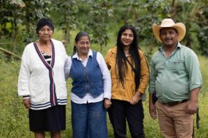 an old woman, a middle-aged woman, a young woman, and a middle-aged man stand smiling in front of vegetation