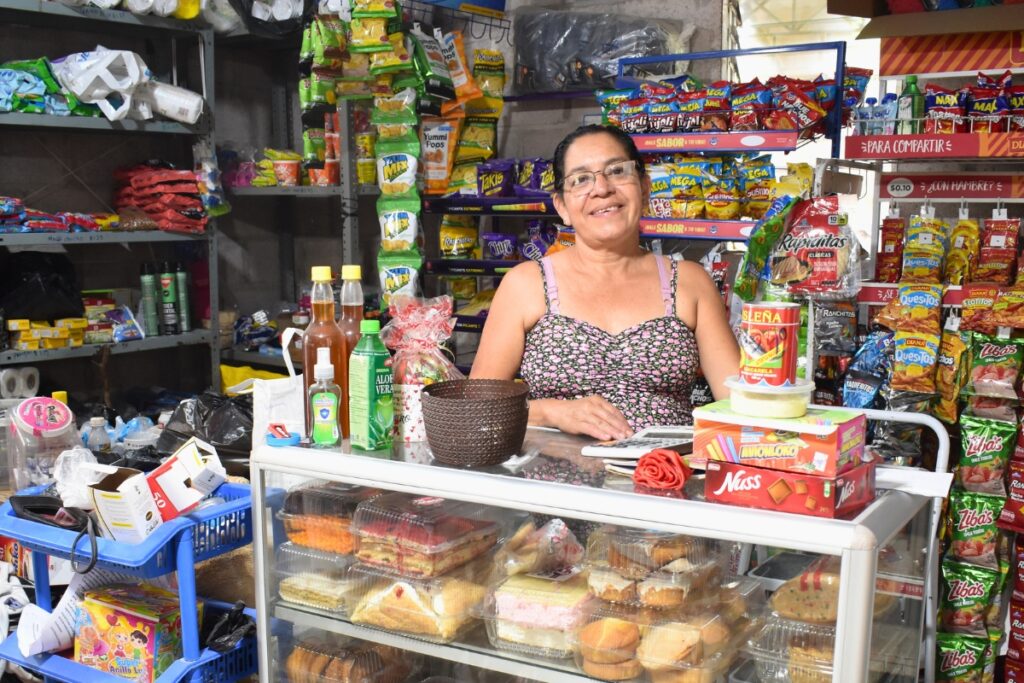 a woman stands smiling behind a counter and in front of shelves with convenience store goods