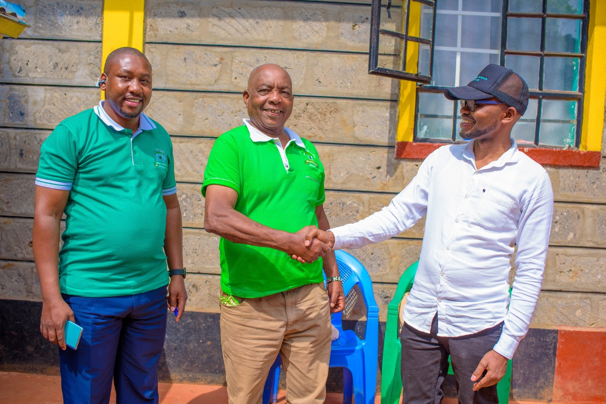 three men stand outside of a building smiling. Two of them are shaking hands