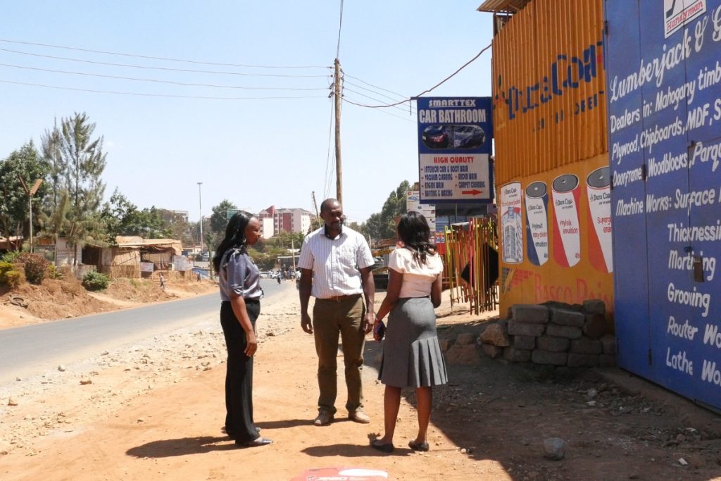 Two women and a man stand on a dirt path in front of a business